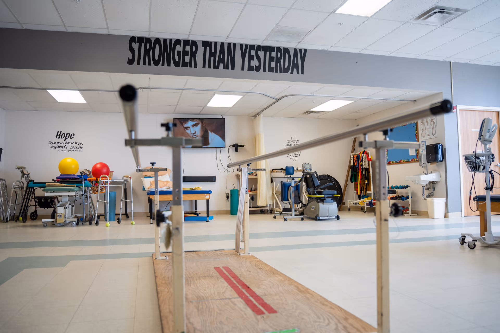 Rehabilitation therapy room with parallel bars in the foreground, exercise equipment, therapy balls, and a 'STRONGER THAN YESTERDAY' sign overhead.
