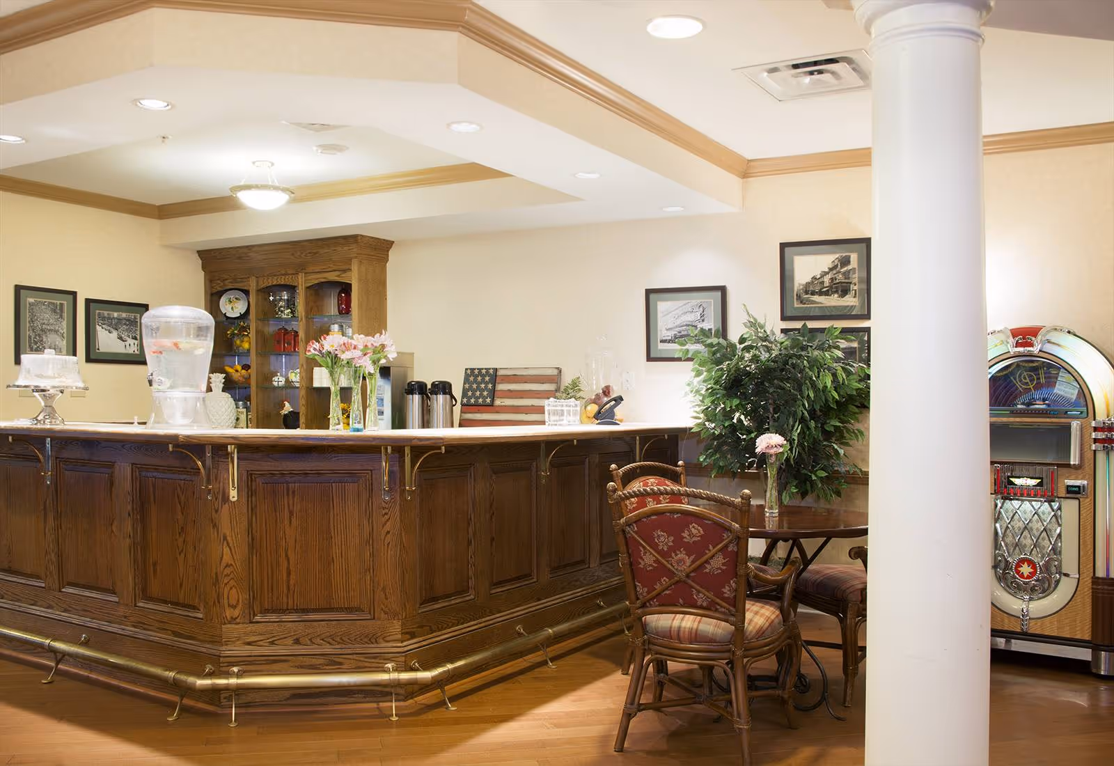 A cozy interior space featuring a wooden bar counter with a water dispenser, coffee pots, and flower vases on top. Behind the bar is a wooden cabinet with decorative items and framed pictures on the wall. In front of the bar, there is a small round table with two cushioned chairs and a potted plant nearby. A vintage jukebox is visible in the corner of the room.