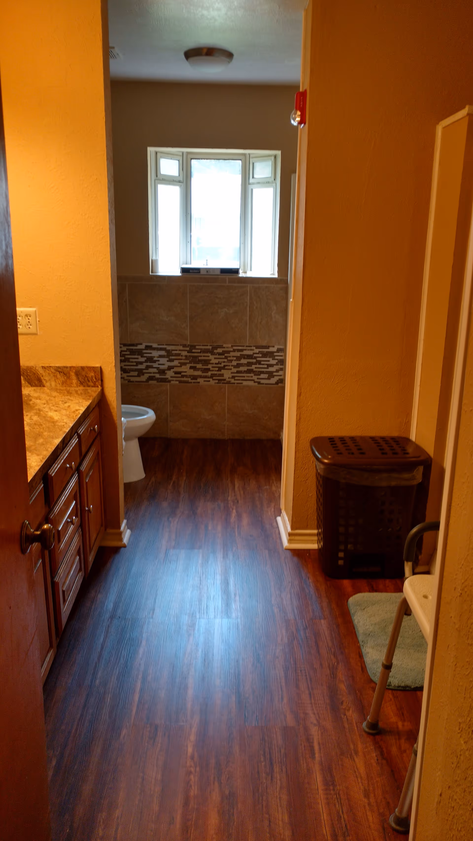 View down a hallway into a bathroom with wood-look flooring, a vanity on the left, a toilet and tiled half-wall with a window ahead, and a laundry basket on the right.