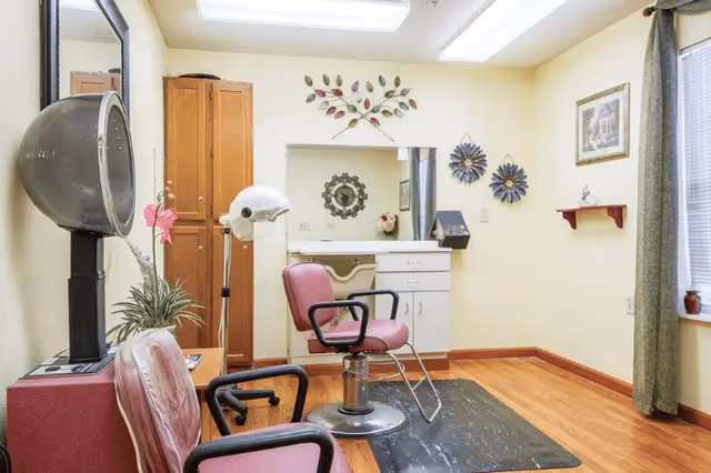 A small salon room with two pink salon chairs, a hair dryer hood, a hair steamer, a wooden cabinet, a white sink with drawers, and decorative wall art. The room has light yellow walls, a window with green curtains, and wooden flooring.