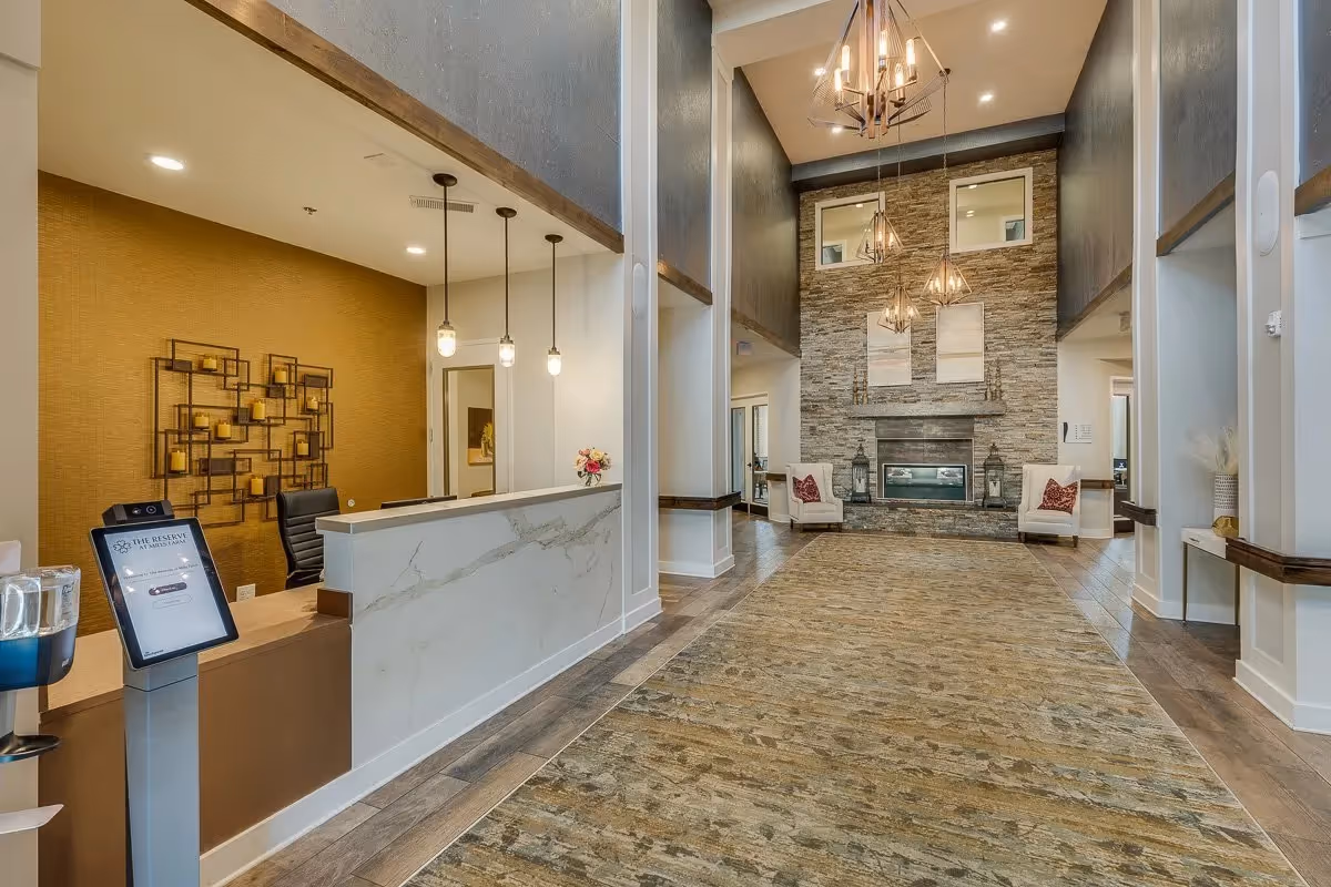 Spacious and well-lit senior living facility lobby with a modern reception desk on the left, featuring a marble front and three hanging pendant lights. The lobby has high ceilings with multiple geometric chandeliers, a large stone fireplace at the far end flanked by two armchairs with red cushions, and a patterned carpet covering the floor.