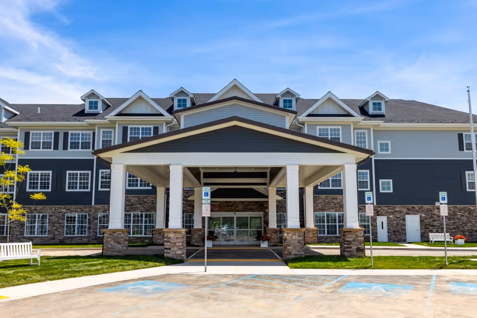 Front exterior view of a large senior living facility building with a covered entrance supported by white columns and stone bases. The building has multiple windows, dormer windows on the roof, and a combination of gray siding and stone facade. There are handicap parking spaces and signs in front of the entrance, along with benches and small trees on the lawn.