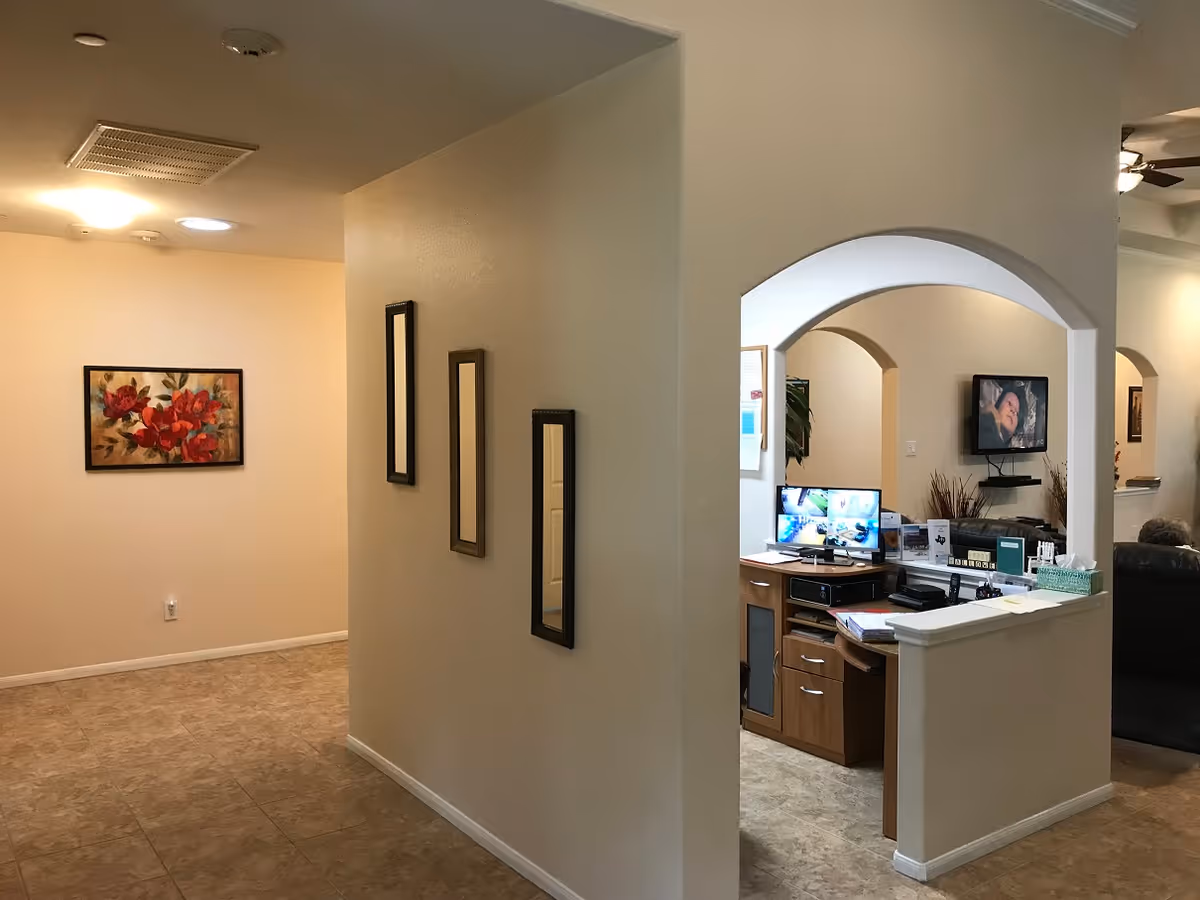 Interior view of a senior living facility showing a hallway with three vertical mirrors on the wall and a framed floral painting. To the right, there is a reception or office area with a wooden desk, computer monitors, and a television mounted on the wall. The floor is tiled and the walls are painted beige.
