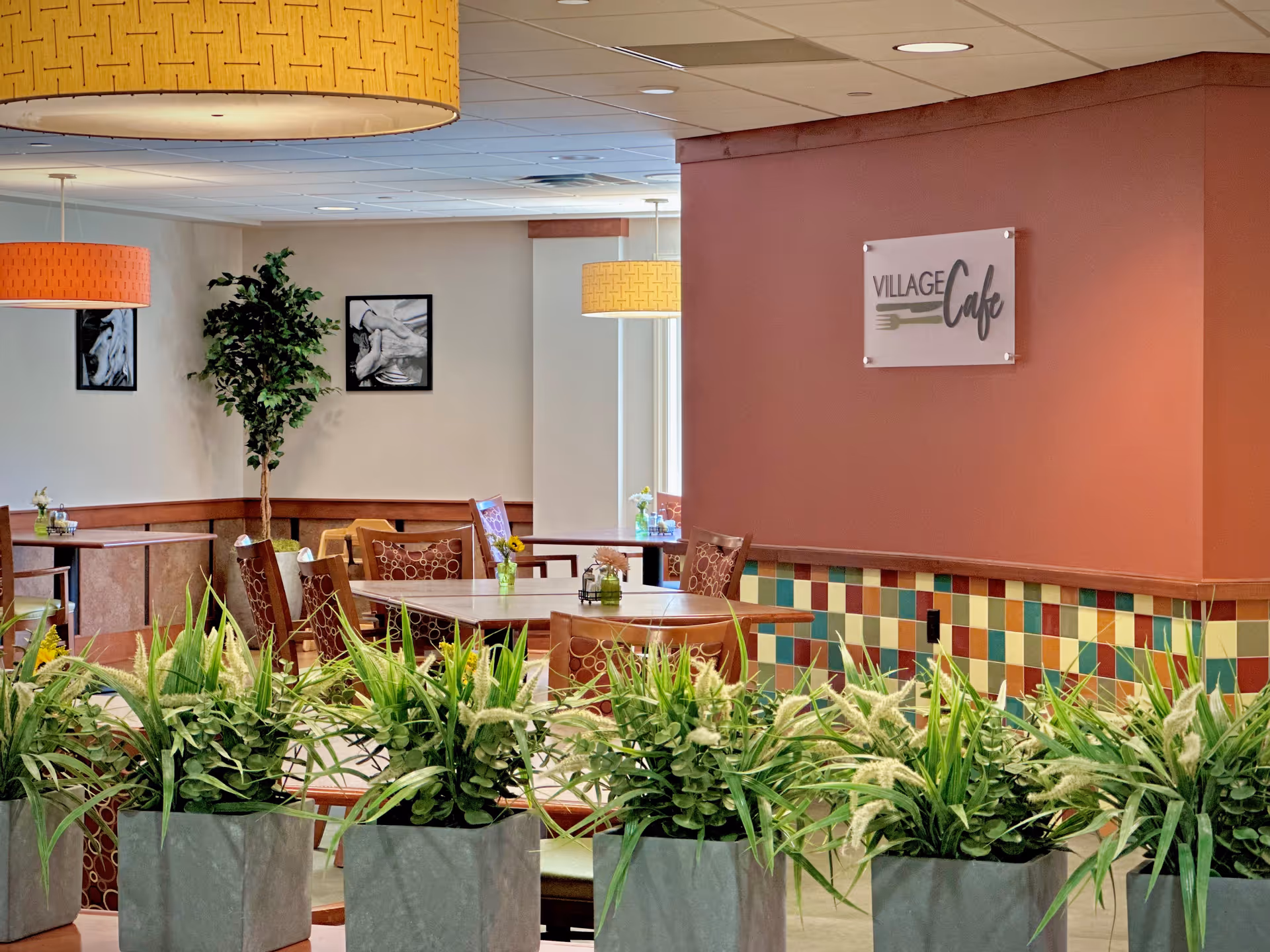 Interior view of a dining area named Village Cafe with several tables and chairs arranged for seating. The space features colorful tiled walls, large hanging light fixtures, framed black and white artwork on the walls, and several green potted plants in the foreground.
