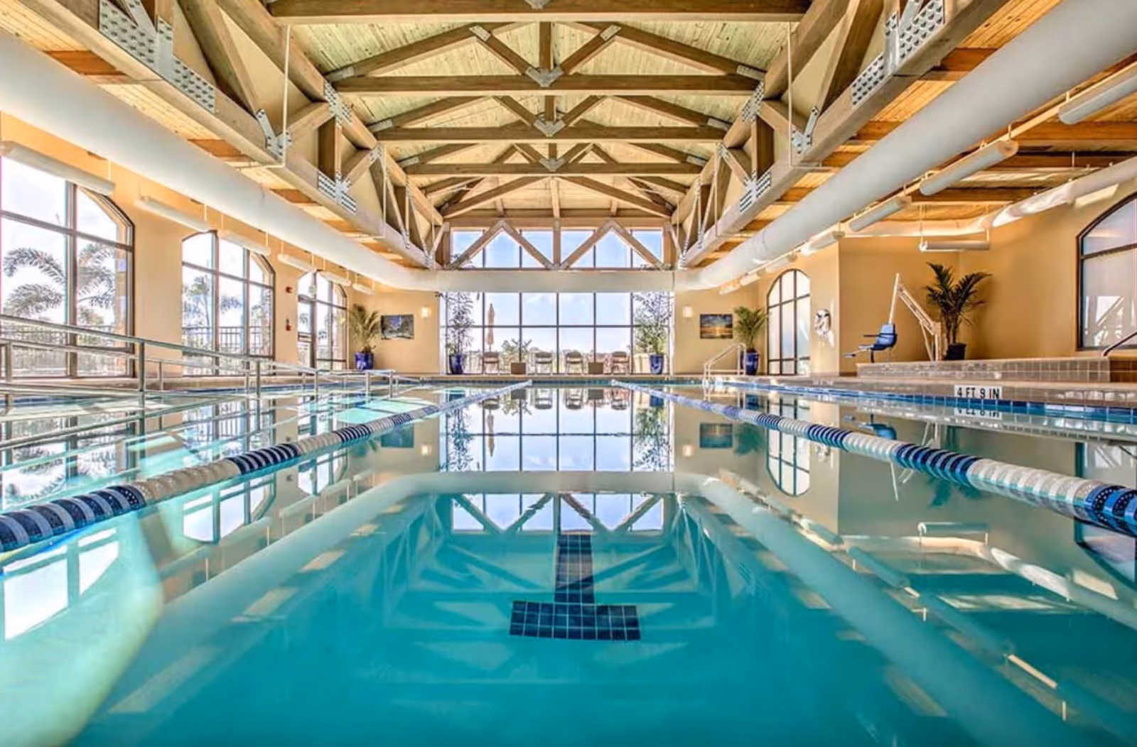 Indoor swimming pool with clear blue water, lane dividers, and a wooden beam ceiling. Large windows on both sides let in natural light and show palm trees outside. There are poolside chairs and potted plants near the windows.