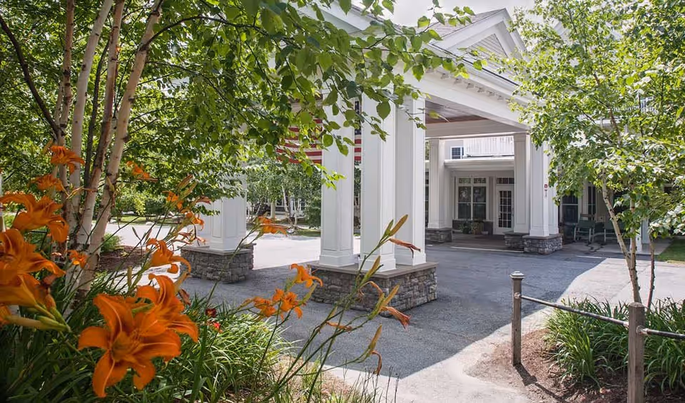 Entrance area of a senior living facility with white columns and stone bases, surrounded by green trees and orange flowers in the foreground.