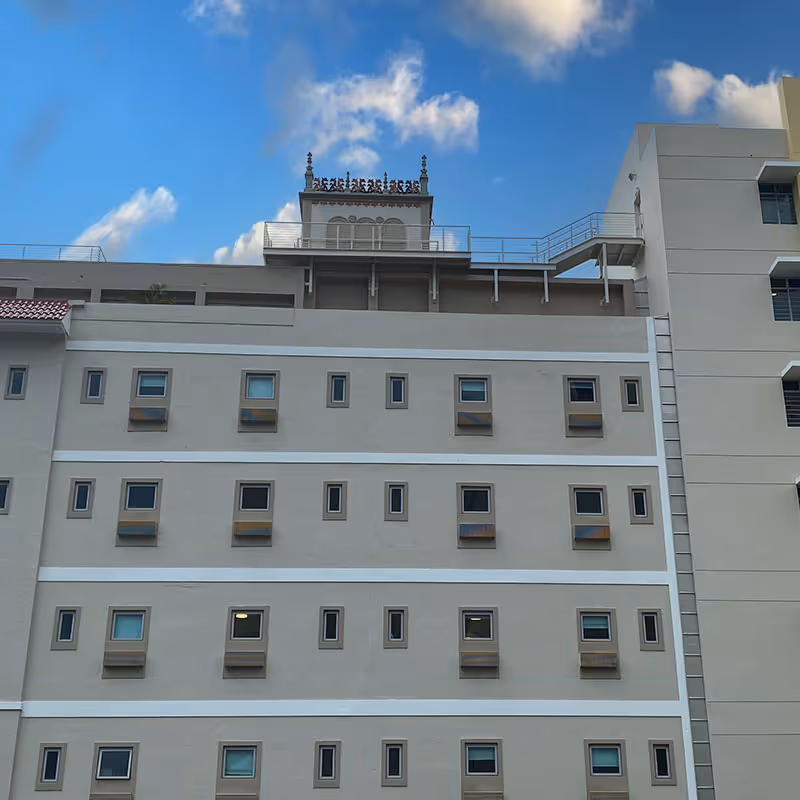 Exterior view of a multi-story building with numerous small rectangular windows arranged in rows. The building has a beige facade with white horizontal bands separating the floors. The top of the building features a decorative structure with ornate ironwork against a blue sky with some clouds.