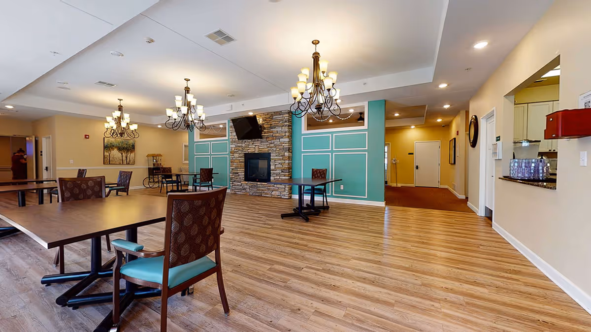 A spacious and well-lit common area in a retirement living facility featuring wooden flooring, multiple tables and chairs, three ornate chandeliers, a stone fireplace with a mounted flat-screen TV above it, and teal accent walls with white trim. There is a hallway leading to other rooms and a counter area on the right side.