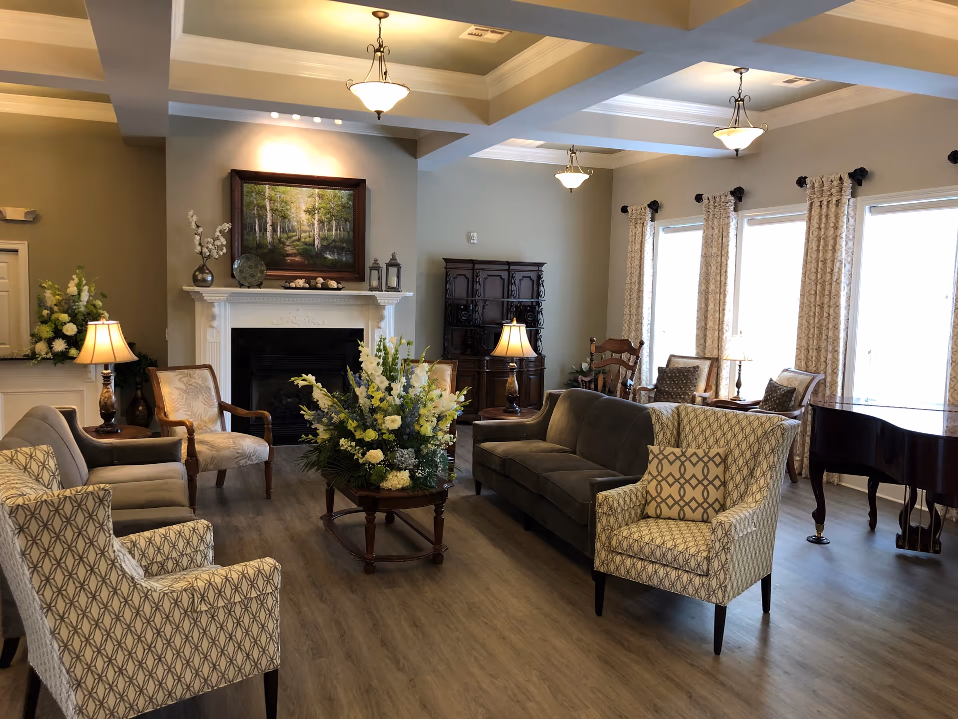 A cozy senior living facility common area with a fireplace, multiple armchairs, a sofa, a wooden coffee table with a large floral arrangement, and a grand piano near large windows with patterned curtains. The room has warm lighting from ceiling fixtures and table lamps, with a painting above the fireplace and wooden furniture along the walls.