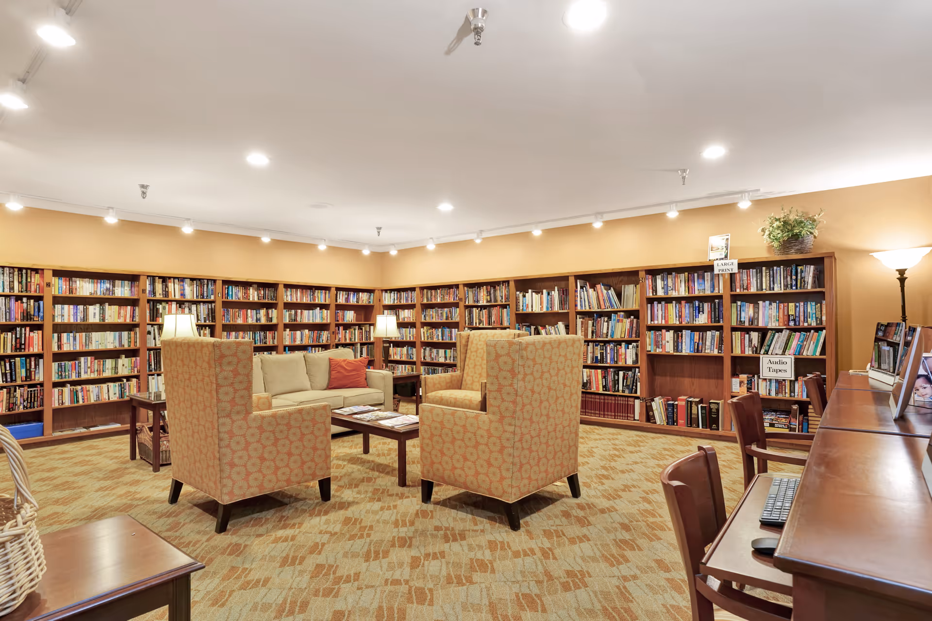 A cozy library room with walls lined with bookshelves filled with books. In the center, there are two patterned armchairs and a beige sofa around a wooden coffee table with magazines. There are table lamps on side tables and a floor lamp providing warm lighting. On the right side, there is a wooden desk with computers and chairs. The room has a carpeted floor and a warm, inviting atmosphere.