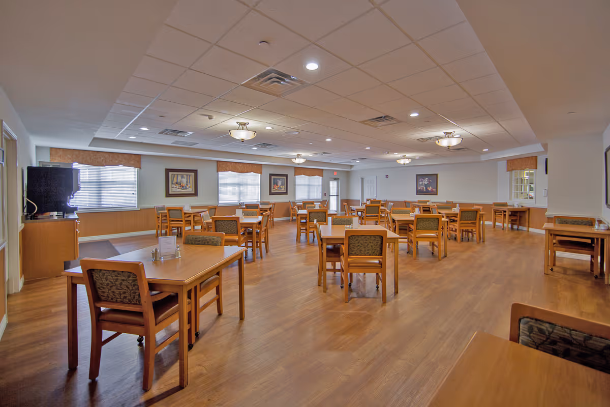 Spacious senior dining room with multiple wooden tables and chairs, hardwood floor, ceiling lights, and windows.