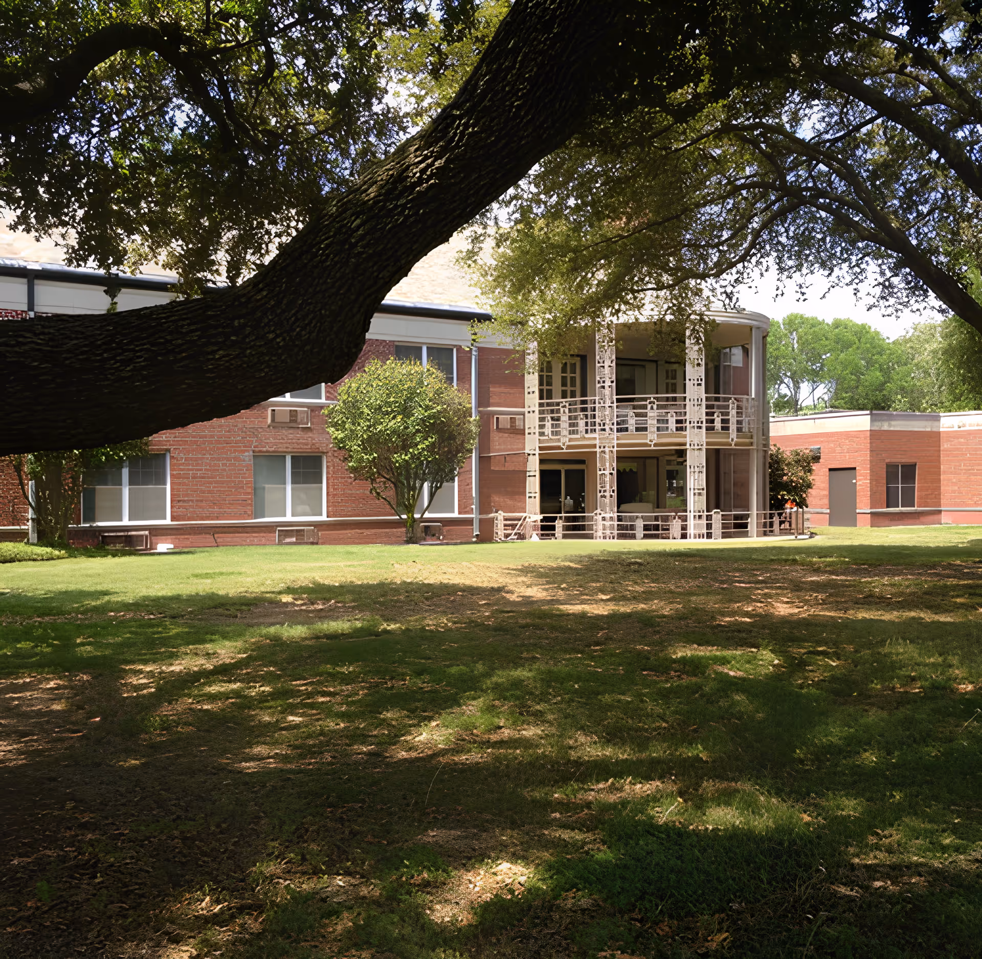 View of a brick building with large windows and a balcony, partially shaded by large tree branches in the foreground, with a grassy lawn in front.