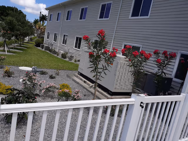 Side yard of a beige multiunit building with a white fence, gravel landscaping, flowering shrubs, and exterior HVAC units.