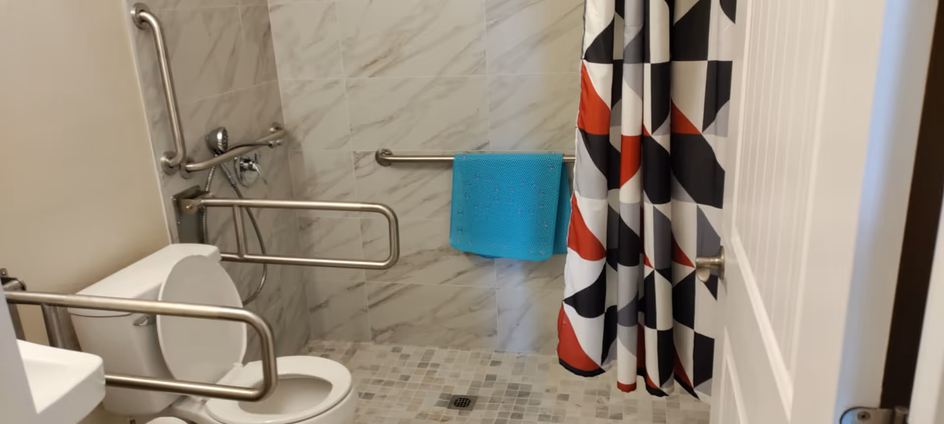 Accessible bathroom featuring a toilet with multiple grab bars, a walk-in tiled shower with a handheld showerhead, a blue towel and a geometric shower curtain.