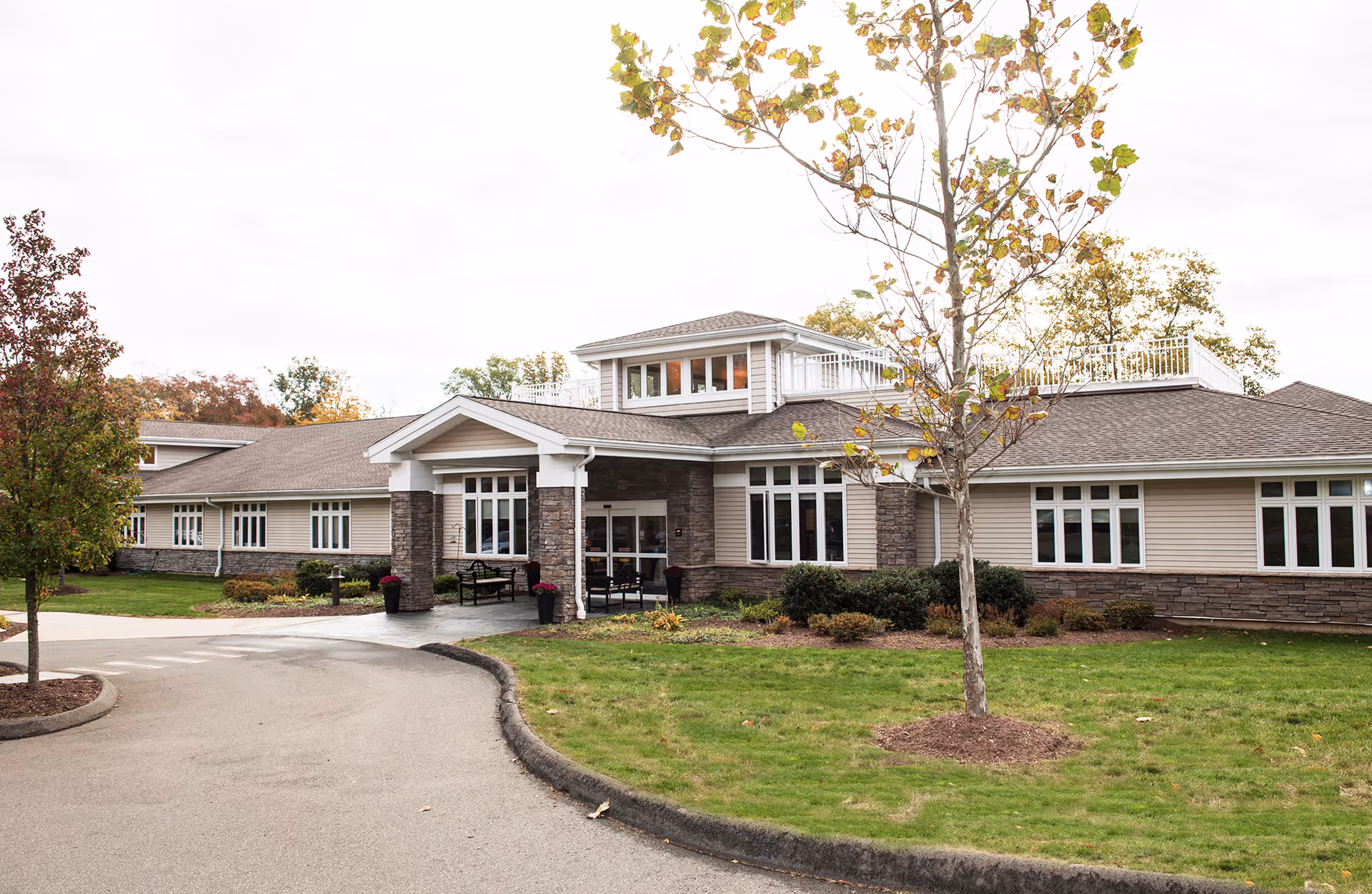 Front exterior of a single-story senior living building with a covered entrance, driveway, lawn, and trees.