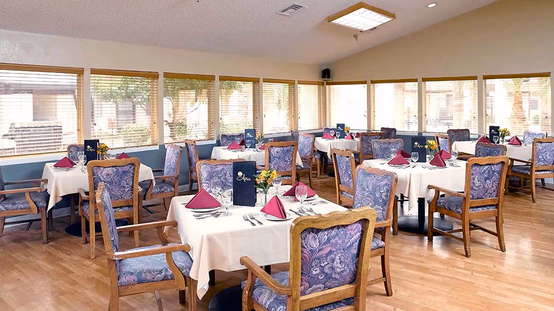 Bright dining room with multiple tables set with white linens, purple napkins, floral-upholstered wooden chairs, and large windows.