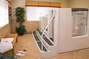 A clean and well-lit room featuring a large white therapeutic bath with steps and handrails for easy access. The room has beige tiled floors and walls, a wooden bench with towels and slippers nearby, a potted plant in the corner, and windows with blinds allowing natural light to enter.