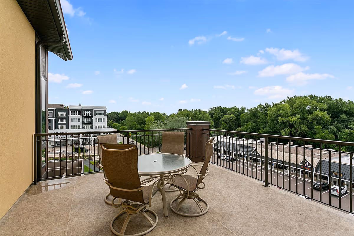 Rooftop balcony with a round glass table and four patio chairs overlooking trees and nearby buildings.