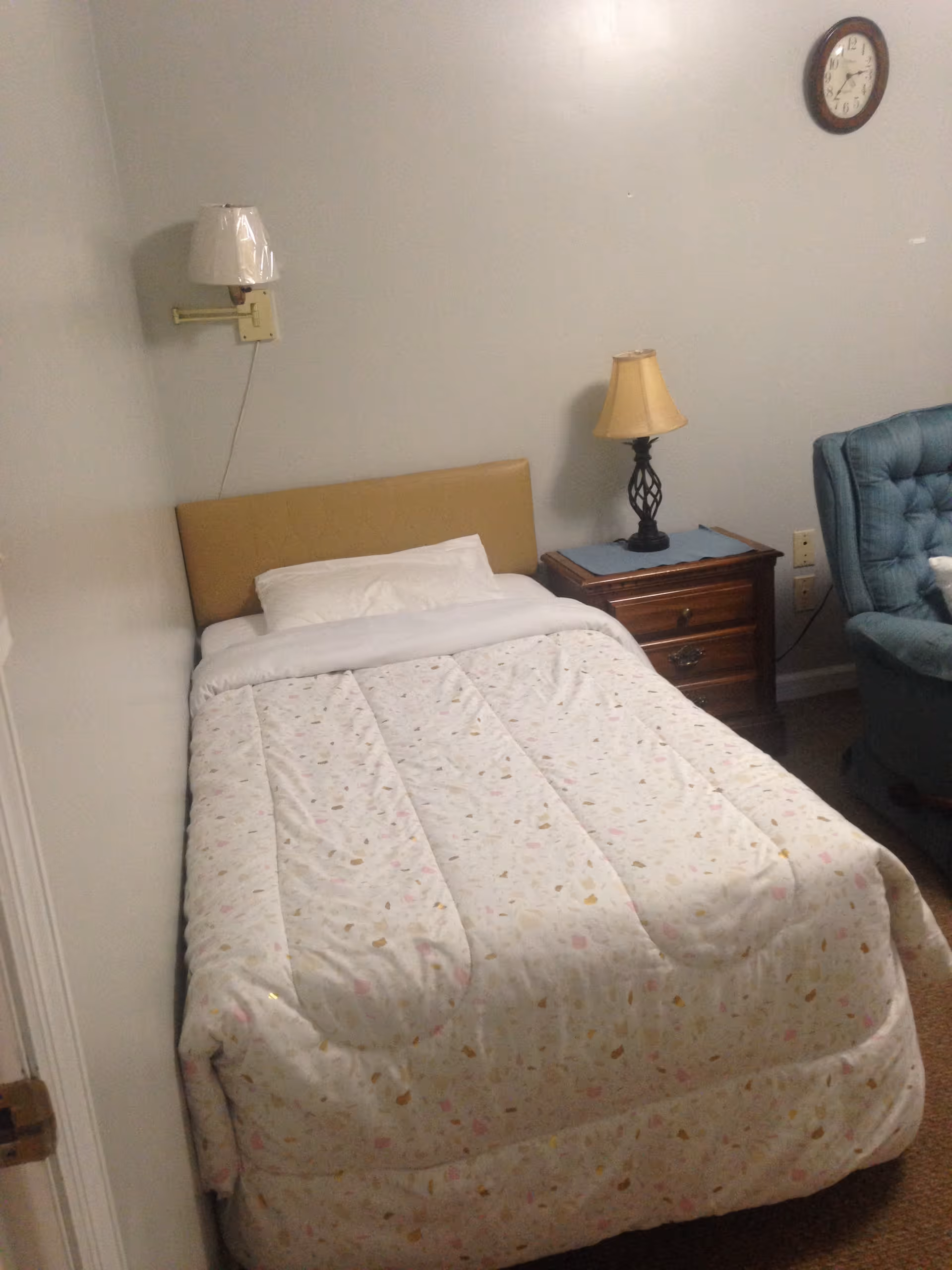 A small bedroom with a single bed covered with a white patterned comforter and a white pillow. There is a wall-mounted lamp above the bed, a wooden nightstand with a table lamp next to the bed, and a blue upholstered armchair in the corner. A round wall clock is visible on the wall above the nightstand.