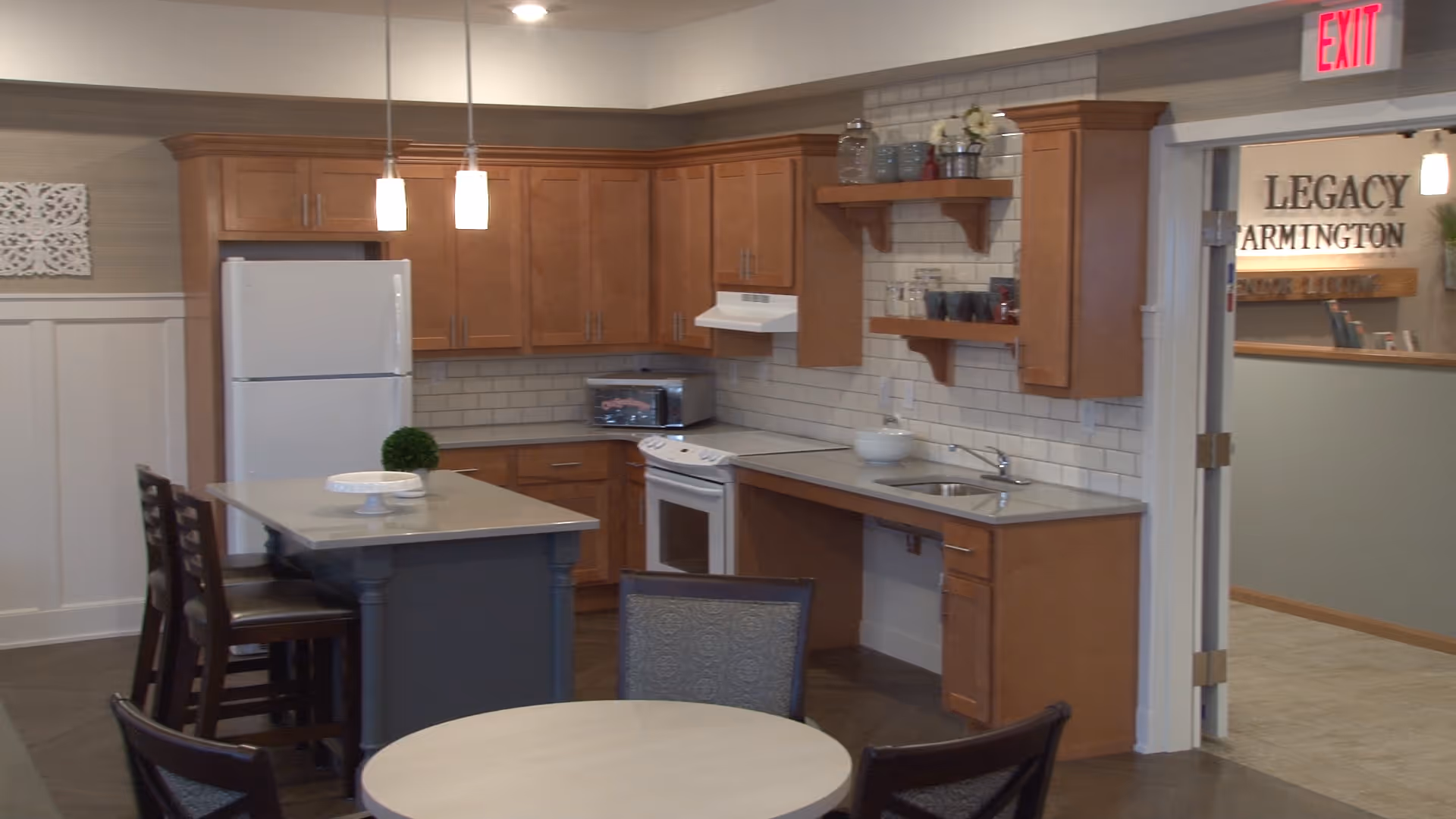 A kitchen area in a senior living facility featuring wooden cabinets, a white refrigerator, a stove with a range hood, a sink, and open shelves with glassware. There is a kitchen island with chairs and a round table with chairs in the foreground. The entrance to another room is visible with a sign that reads 'Legacy of Farmington Senior Living'.
