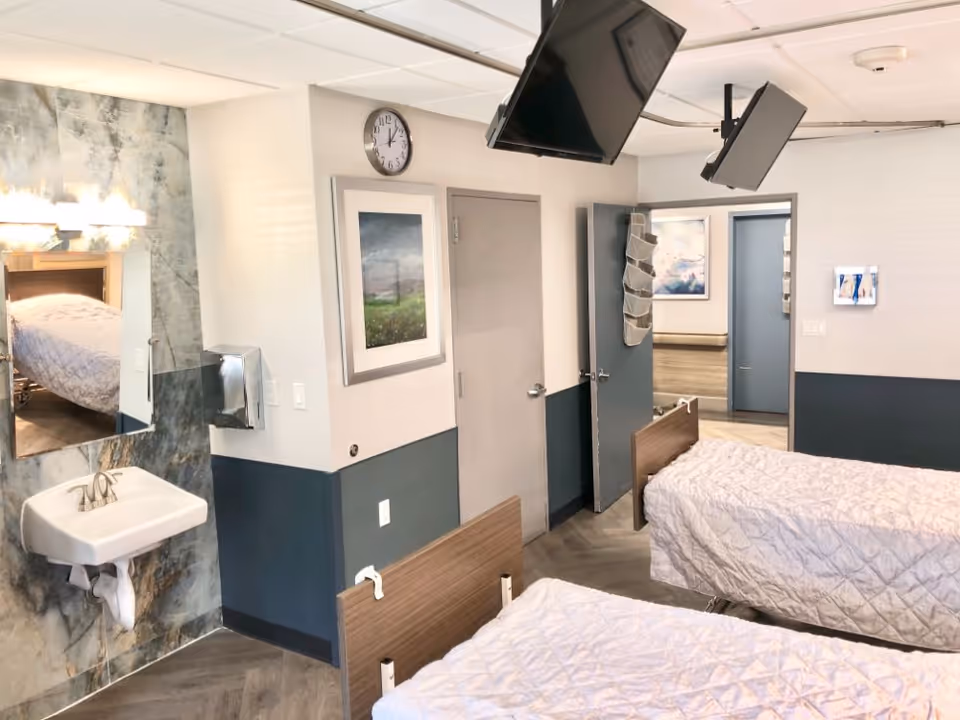 A room with two hospital-style beds covered with white quilts, a wall-mounted sink with a mirror above it, two wall-mounted televisions, a clock, and two doors leading to other rooms. The walls are painted white and dark blue with framed pictures hanging on them.
