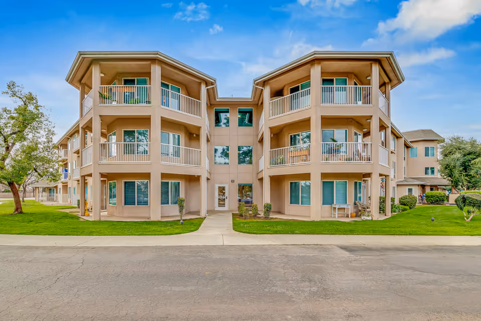 Exterior view of a three-story senior living facility building with beige walls, multiple balconies, and large windows. The building is surrounded by green grass, a few trees, and a paved driveway in front under a partly cloudy blue sky.