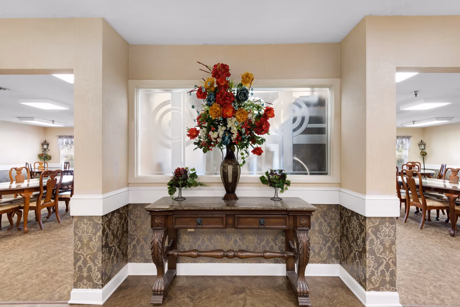 A decorative wooden table with a marble top is placed against a wall with patterned wallpaper. On the table is a large vase filled with colorful artificial flowers in red, yellow, and blue, flanked by two smaller decorative plants. On either side of the table, there are openings leading to dining areas with wooden chairs and tables, and windows with floral curtains.