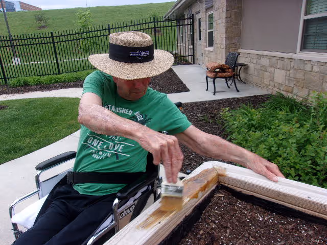 A man in a wheelchair wearing a straw hat and green t-shirt is painting the wooden edge of a raised garden bed outdoors next to a stone building with a black metal fence and green grass in the background.