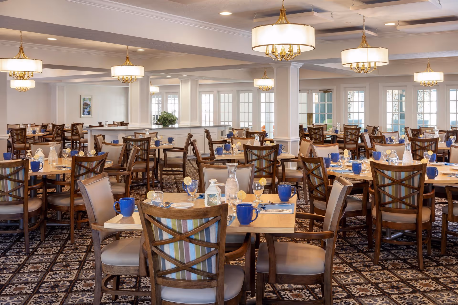 A spacious dining room with multiple wooden tables and chairs arranged neatly. Each table is set with blue mugs, glasses with lemon slices, plates, and silverware. The room features patterned carpet flooring, white walls, large windows with glass doors, and elegant ceiling lights with gold accents.