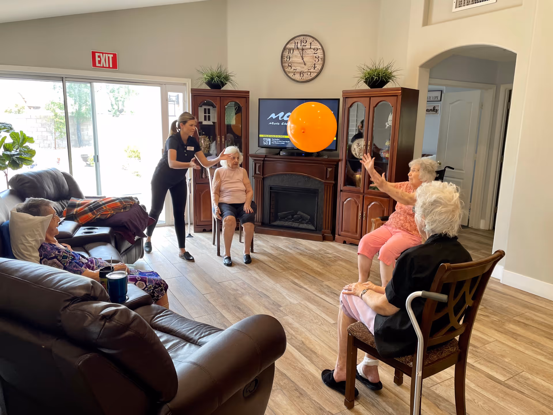 A group of elderly women seated in a living room area playing with a large orange balloon. A caregiver is standing and assisting one of the women. The room has wooden flooring, a large clock on the wall, a TV above a fireplace, and glass cabinets on either side. There is a sliding glass door with an exit sign above it letting in natural light.