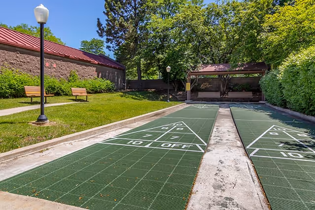 Outdoor shuffleboard courts with green playing surfaces and white markings, surrounded by grass, benches, lamp posts, trees, and a building with a red roof under a clear blue sky.
