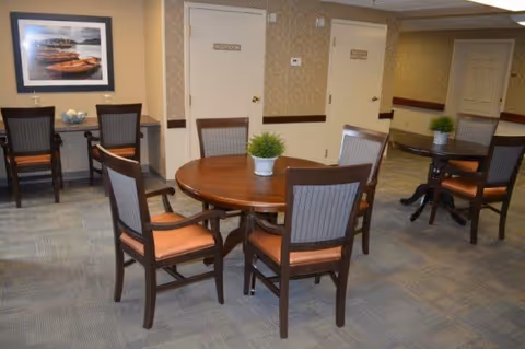 A seating area with round wooden tables, chairs, and small potted plants in an assisted living facility interior.