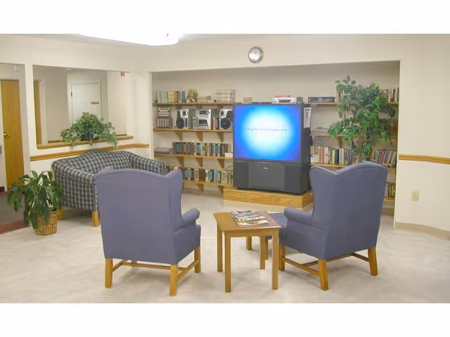 A community lounge with two blue armchairs and a small table facing a television and bookshelf, with a couch and plants in the background.