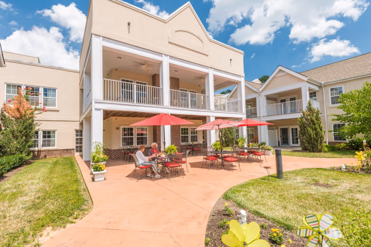 Outdoor patio area at Park Place of West Knoxville with tables and chairs under red umbrellas. Two elderly women are sitting and conversing at one of the tables. The building has a beige exterior with white railings on the balconies and a clear blue sky with some clouds above.