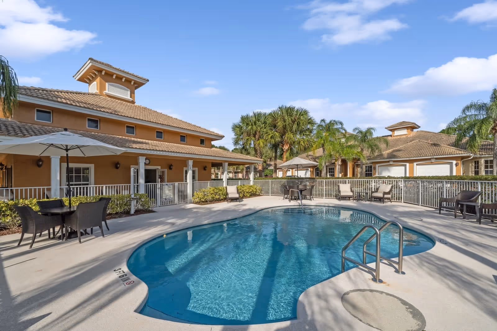 Outdoor swimming pool area at The Palms at St. Lucie West with clear blue water, surrounded by a concrete deck with lounge chairs, tables with umbrellas, and a fence. The background shows tan-colored buildings with tiled roofs and palm trees under a blue sky with some clouds.