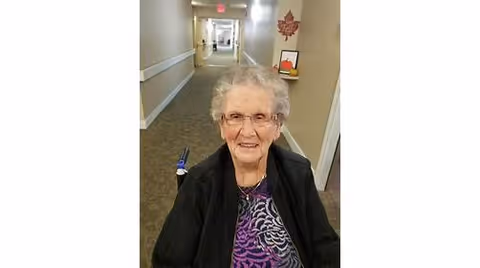 An elderly woman with short gray hair and glasses is smiling while sitting in a wheelchair in a hallway of a senior living facility. The hallway has beige walls, a carpeted floor, handrails on the walls, and a decorative maple leaf wall hanging in the background.