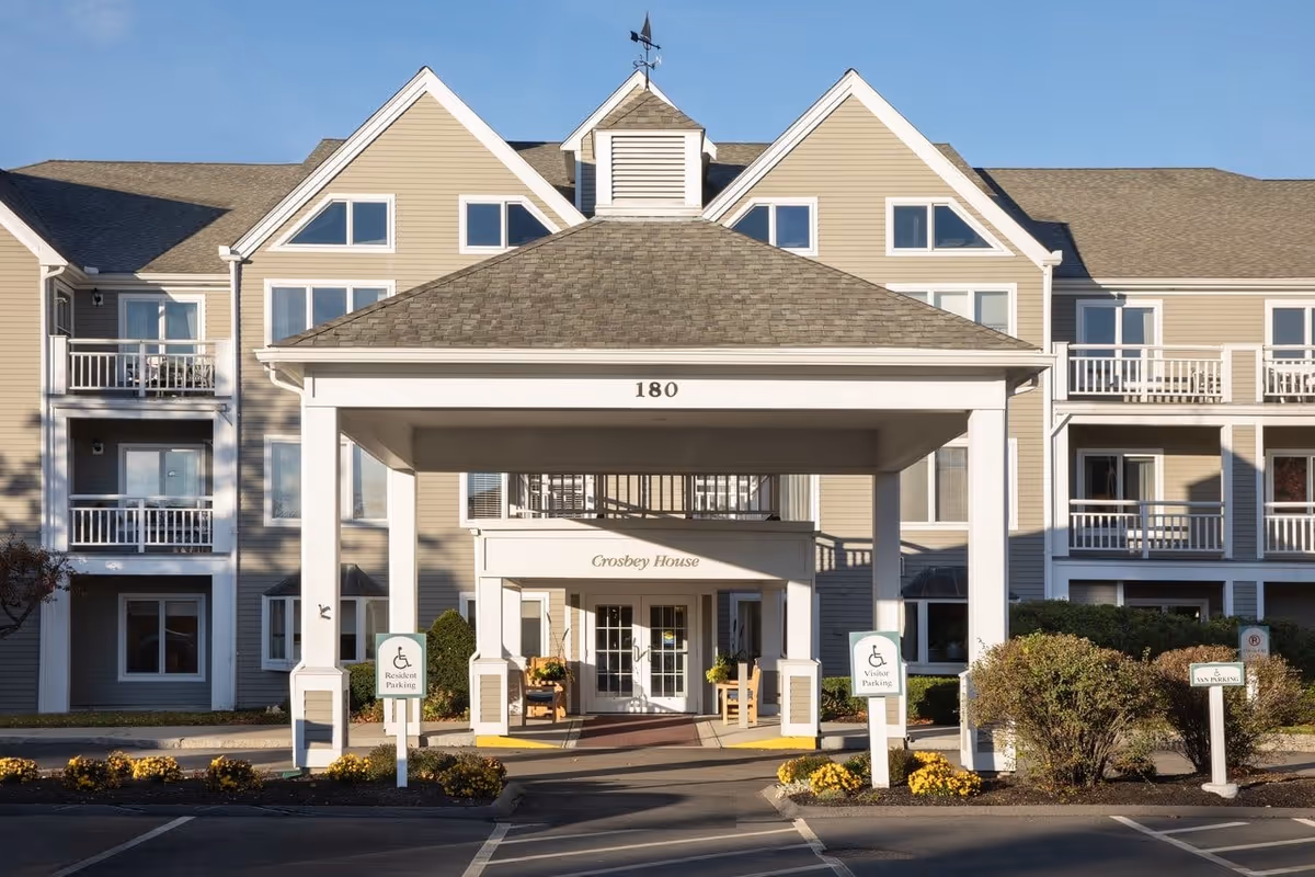 Front exterior view of a senior living facility named Crosbey House at 180, featuring a covered entrance with white pillars, balconies, and multiple windows under a clear blue sky. There are designated parking signs for resident and visitor parking near the entrance.
