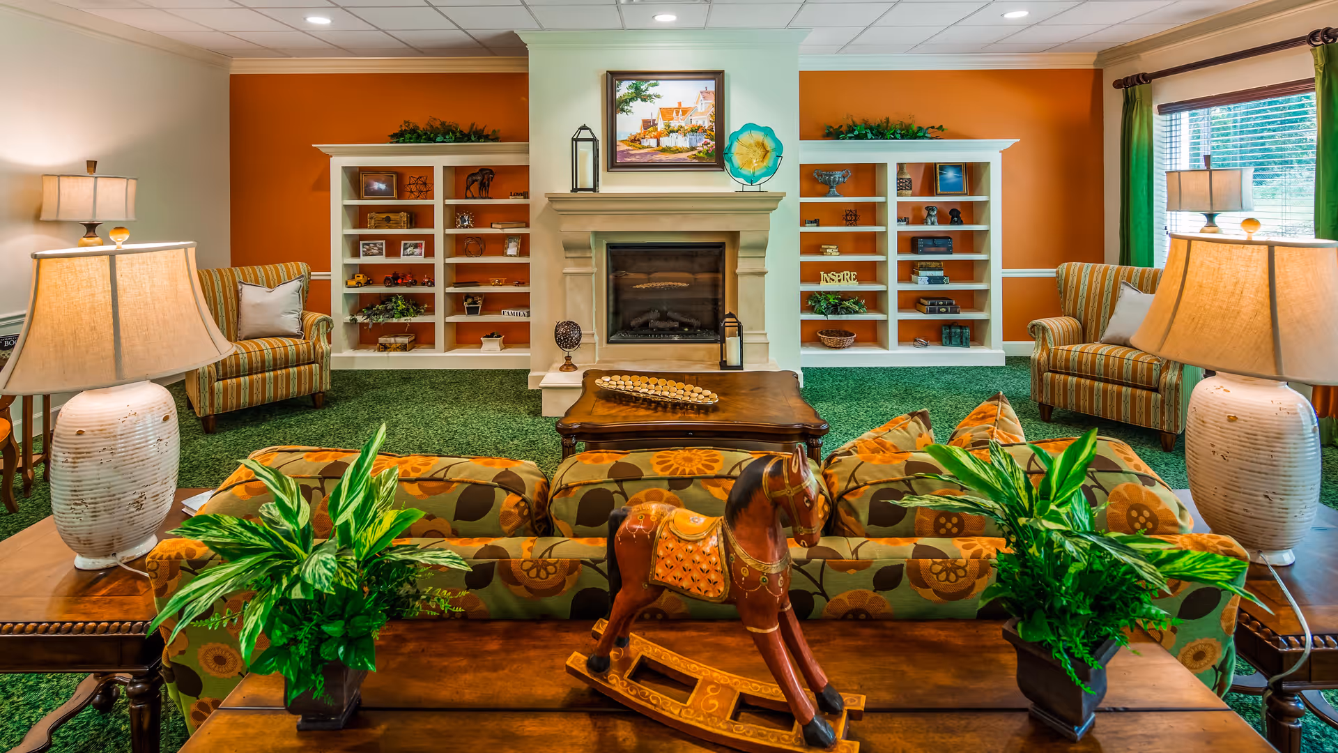 A cozy living room with a green carpet, floral patterned sofa, two striped armchairs, and a wooden coffee table. The room features a fireplace with built-in white shelves on either side, decorated with various ornaments and plants. Two table lamps with beige shades are placed on side tables next to the sofa. A wooden rocking horse and two potted plants are on a table behind the sofa. A window with green curtains allows natural light into the room.