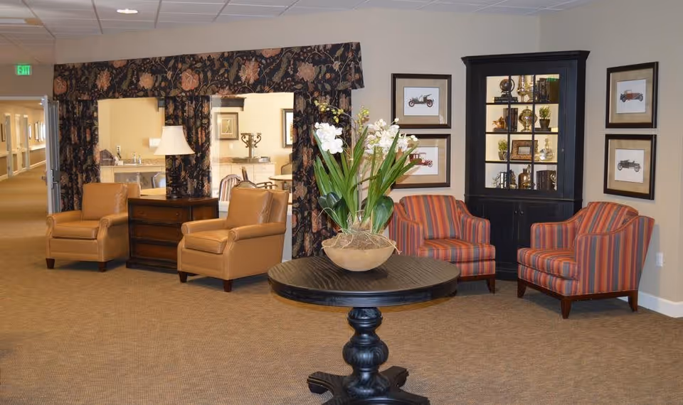 A cozy sitting area in an assisted living facility featuring two tan leather armchairs with a wooden side table and lamp between them, a round black pedestal table with a large floral arrangement in the center, and two striped armchairs next to a black display cabinet filled with decorative items. The walls are adorned with framed vintage car prints, and a hallway is visible in the background.