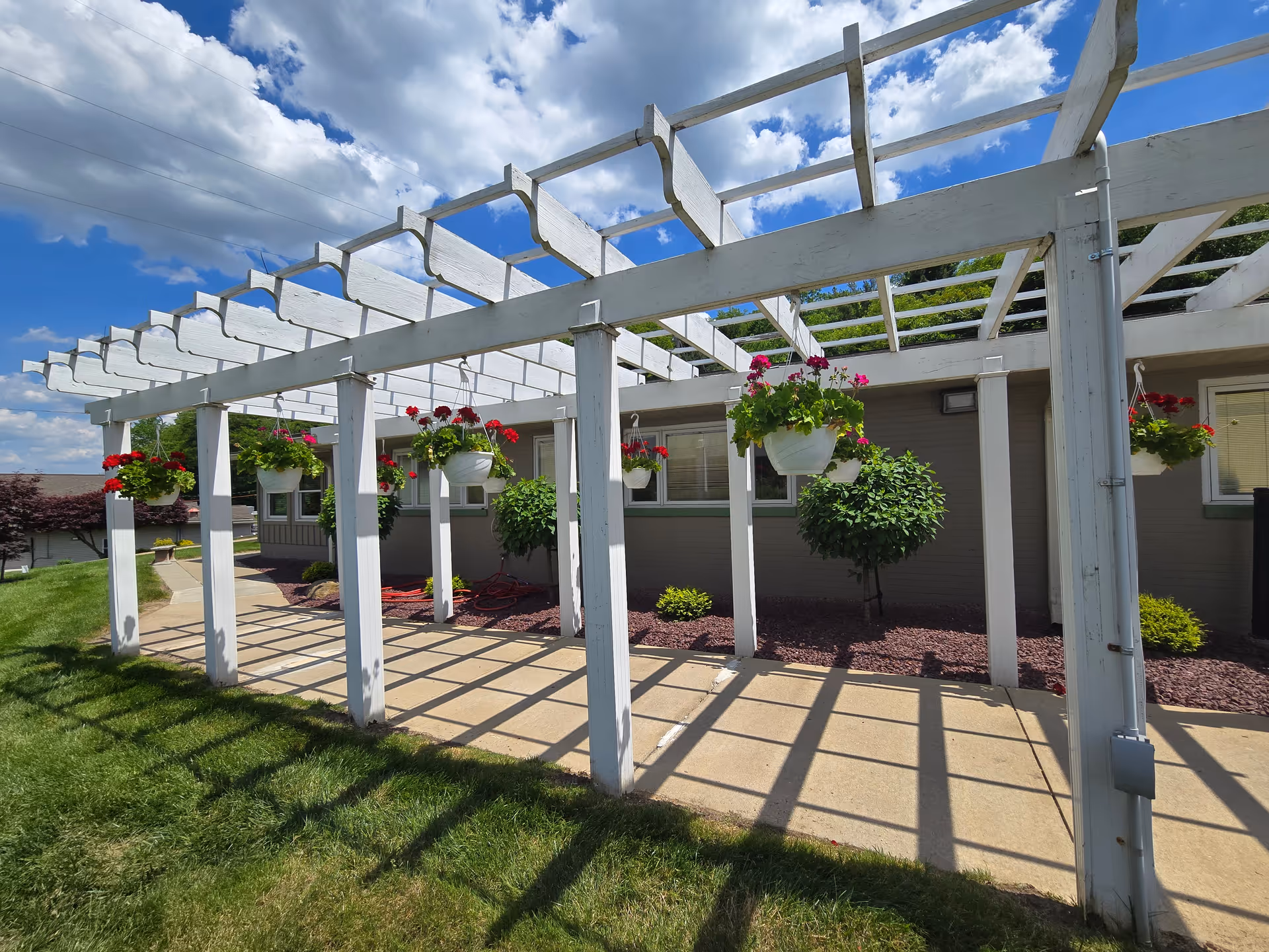White wooden pergola with hanging flower baskets along a walkway beside a single-story building under a blue sky.
