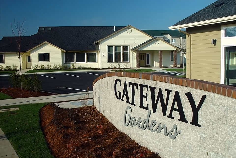 Exterior view of the Gateway Gardens facility showing a large stone sign with the facility name in front of a beige building with multiple windows and a dark roof under a clear blue sky.