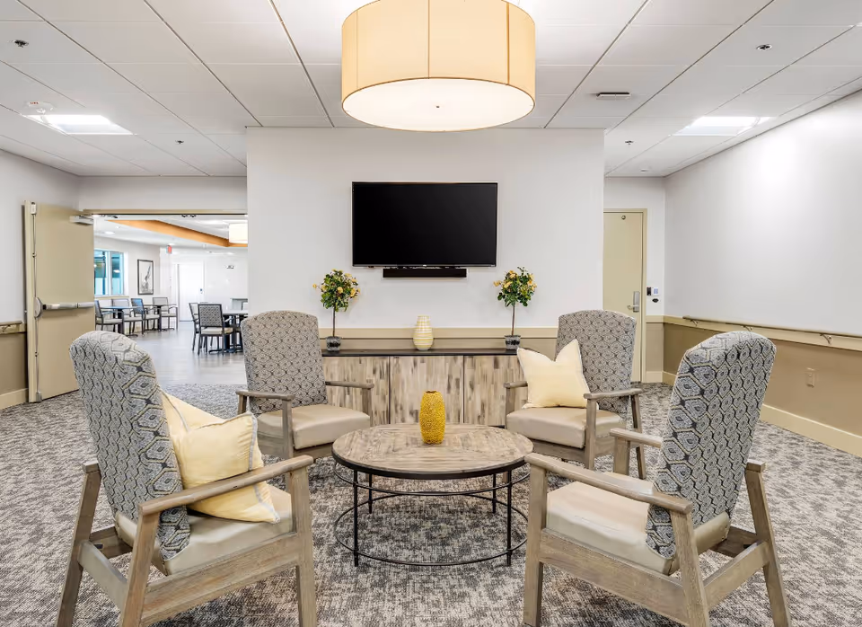 A cozy senior living common area with four patterned armchairs arranged around a round wooden coffee table. The chairs have light cushions and some have yellow throw pillows. A wooden cabinet against the wall holds two small potted plants and a decorative vase. Above the cabinet is a flat-screen TV mounted on the wall. The room has a neutral color palette with beige and gray tones, carpeted floor, and a large ceiling light fixture. In the background, there is an open doorway leading to a dining area with tables and chairs.