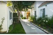 Sunlit outdoor walkway between white senior living buildings with trees and landscaped greenery