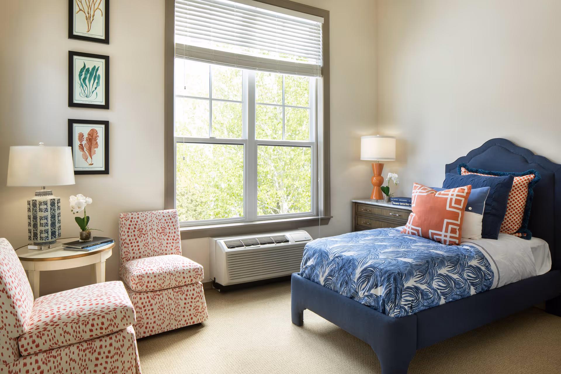 A bright bedroom with a large window letting in natural light. The room features a blue upholstered bed with multiple decorative pillows in blue, white, and orange patterns. There are two patterned chairs with red and white spots next to a small round table holding a lamp and a small plant. On the wall beside the window are three framed botanical prints. A wooden nightstand with a lamp and a small plant is beside the bed.