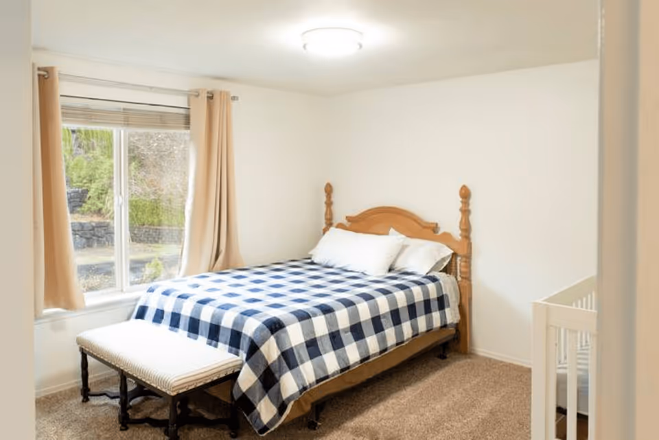 A bedroom with a wooden bed frame, a blue and white checkered blanket, two white pillows, beige curtains on a window, a small bench at the foot of the bed, and a white crib in the corner.