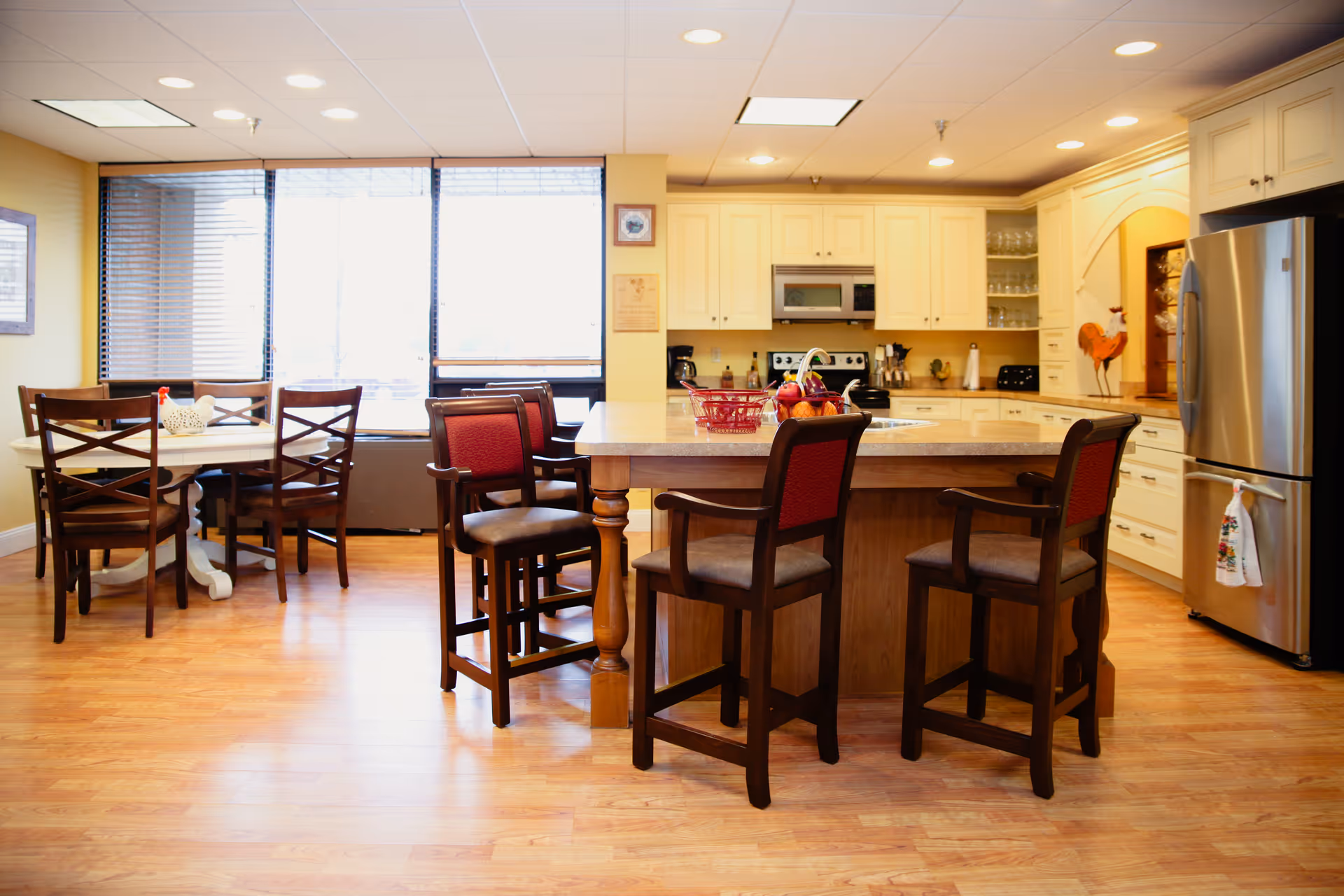 A bright and spacious kitchen and dining area with wooden floors, a large kitchen island with four high chairs, white cabinetry, stainless steel refrigerator, microwave, and stove. There is a round dining table with four wooden chairs near a large window with blinds.