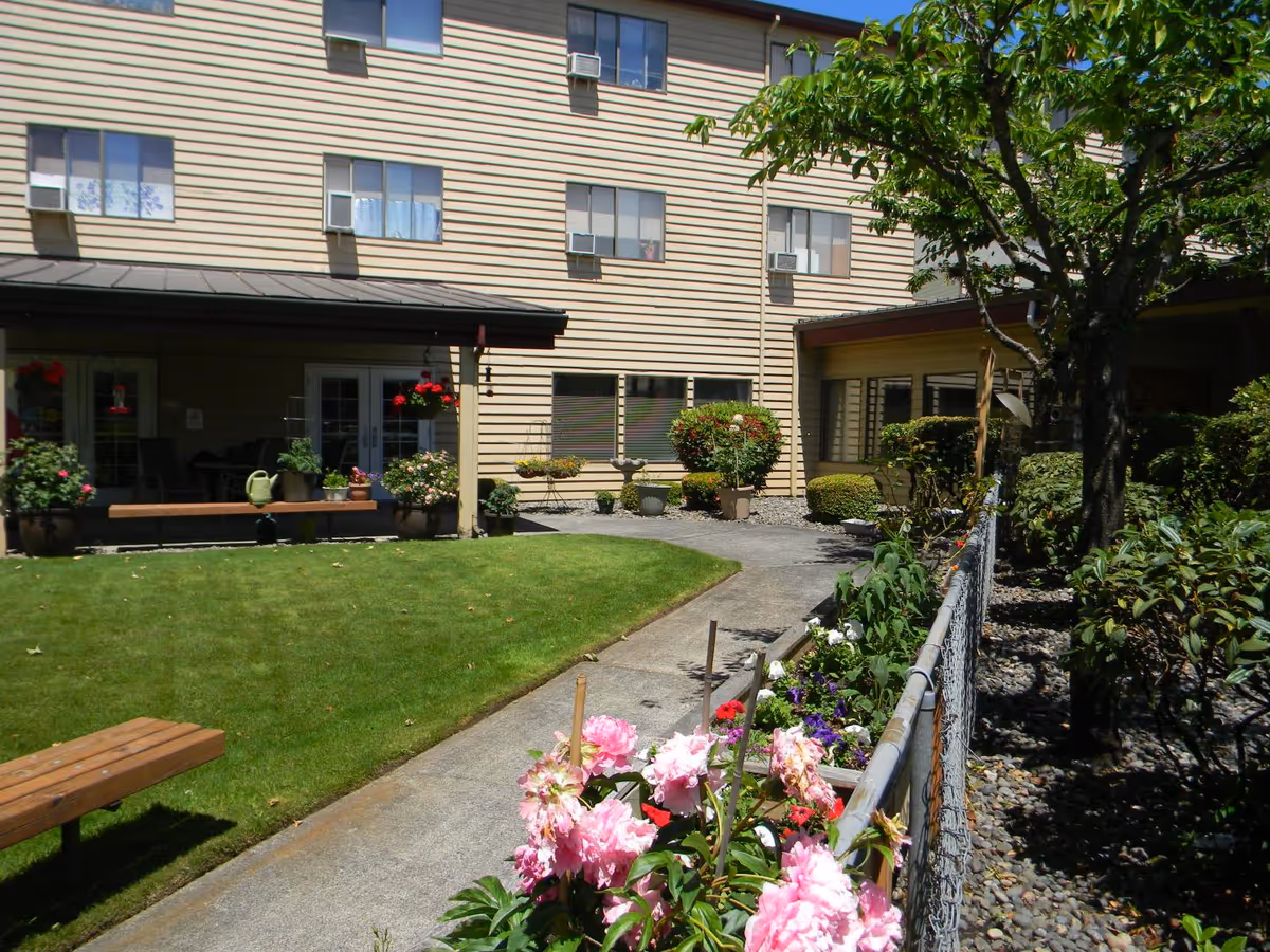 Outdoor courtyard area of a senior living facility with a green lawn, a concrete walkway, flower beds with pink and purple flowers, potted plants, and a beige multi-story building with windows and air conditioning units in the background.