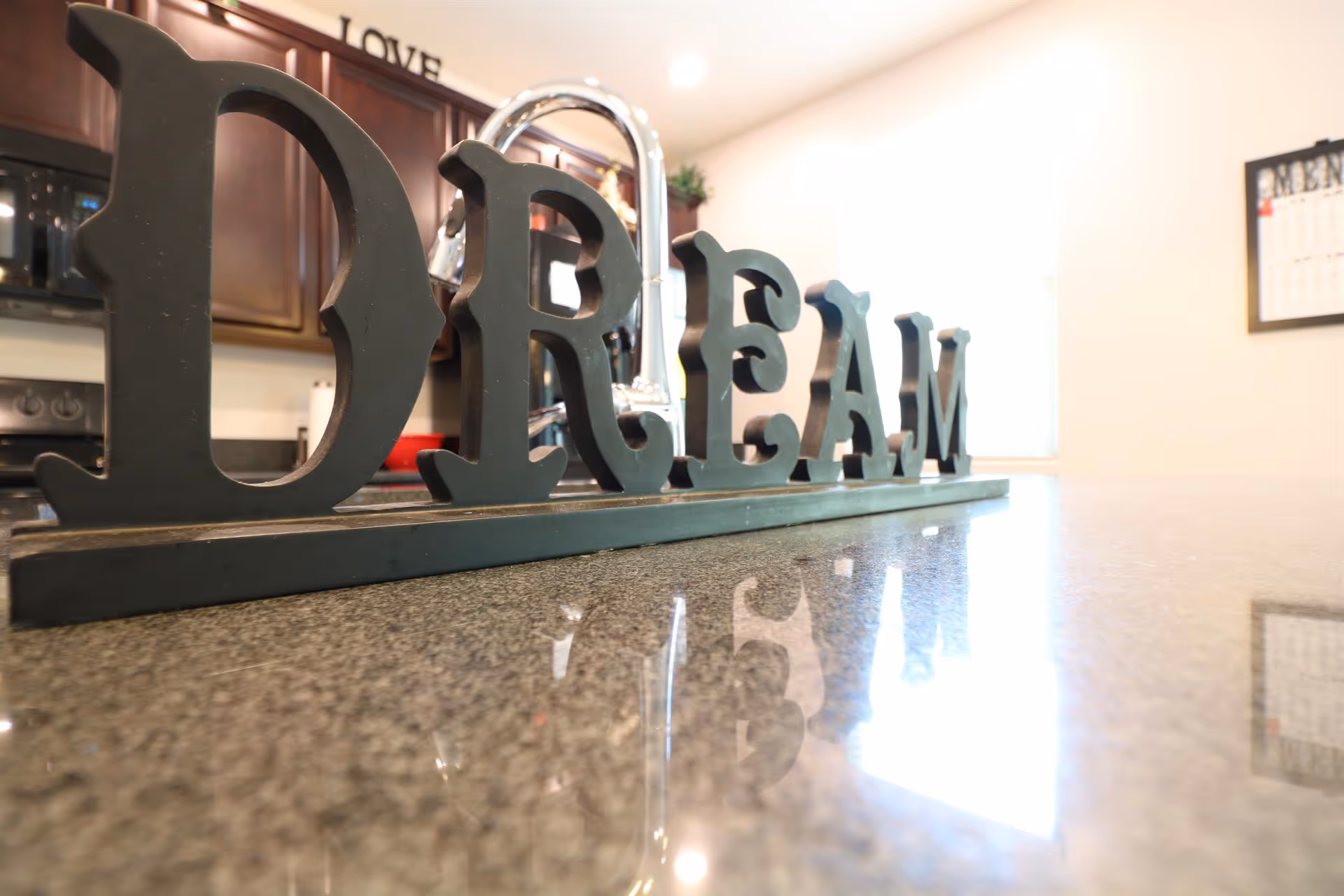 Close-up view of a decorative sign spelling the word 'DREAM' placed on a kitchen countertop with a shiny granite surface. In the background, there are dark wooden cabinets, a stove, and a calendar on the wall.