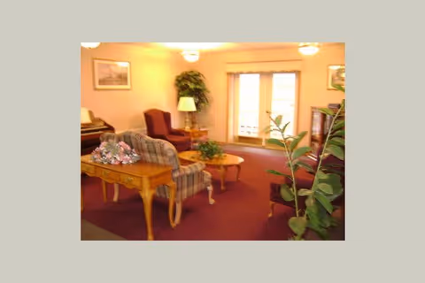 A cozy living room area with a plaid upholstered armchair, a wooden side table with a floral arrangement, a round wooden coffee table, a burgundy armchair, a floor lamp, potted plants, framed pictures on the walls, and large windows with curtains letting in natural light.
