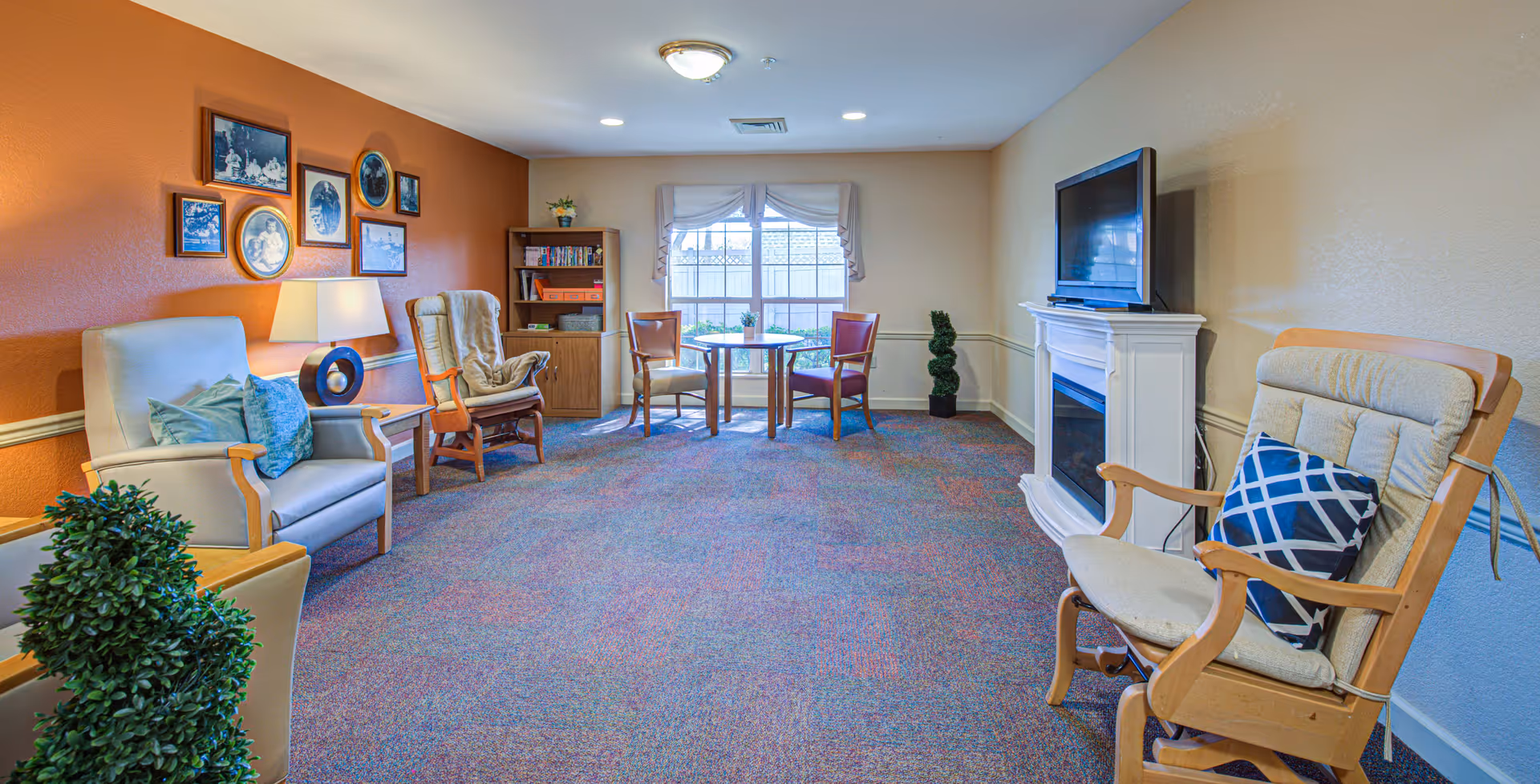 A cozy living room with multiple armchairs, a small round table with two chairs near a window, a white electric fireplace with a TV on top, a bookshelf, and framed photos on an orange accent wall. The room has carpeted flooring and soft lighting.